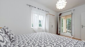 Ein Schlafzimmer mit einem gemusterten Bettbezug, einem Fenster mit transparenten Vorhängen und Blick auf Grün.