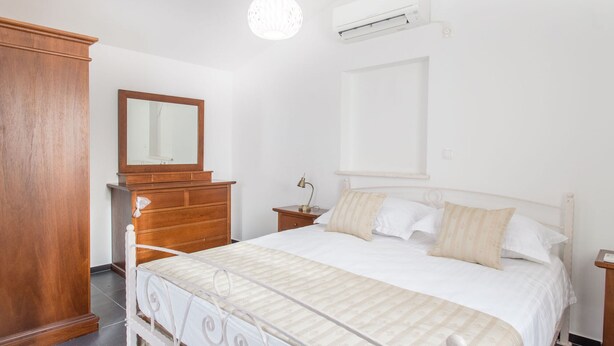 Ein Schlafzimmer mit Bett, Holzkommode, Spiegel, Nachttisch, Lampe und einer Klimaanlage.