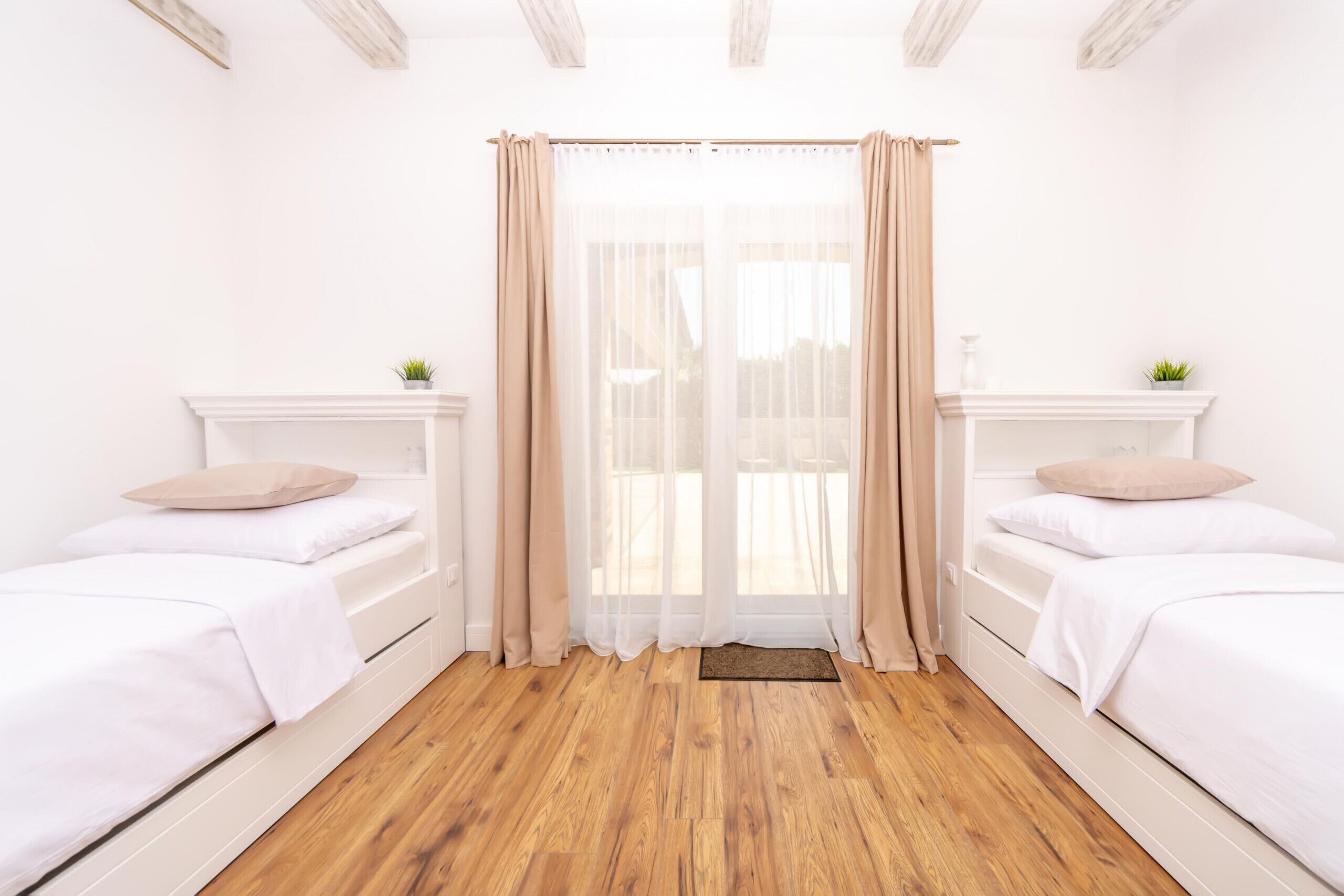 A room with two single beds, a wooden floor, white walls, and a window with sheer curtains.
