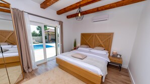 A bedroom with a wooden bed, a nightstand, a mirror, and a view of an outdoor swimming pool. - Villa Idassa (Policnik)