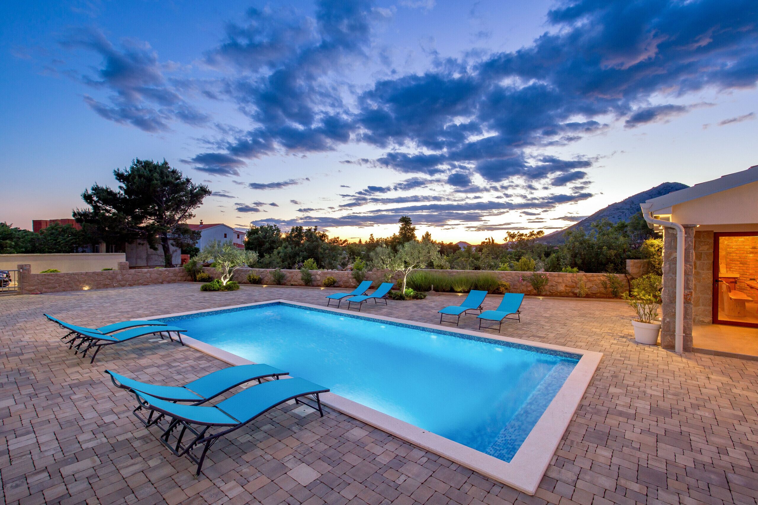 A rectangular swimming pool with a tiled edge, surrounded by a brick patio. There are multiple lounge chairs aligned along the pool's edge.