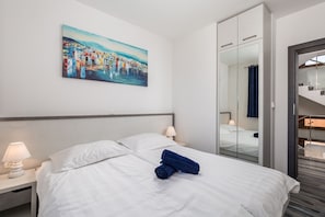 A hotel room with a bed, white bedding, a painting on the wall, a nightstand with a lamp, and a mirror in the closet.
