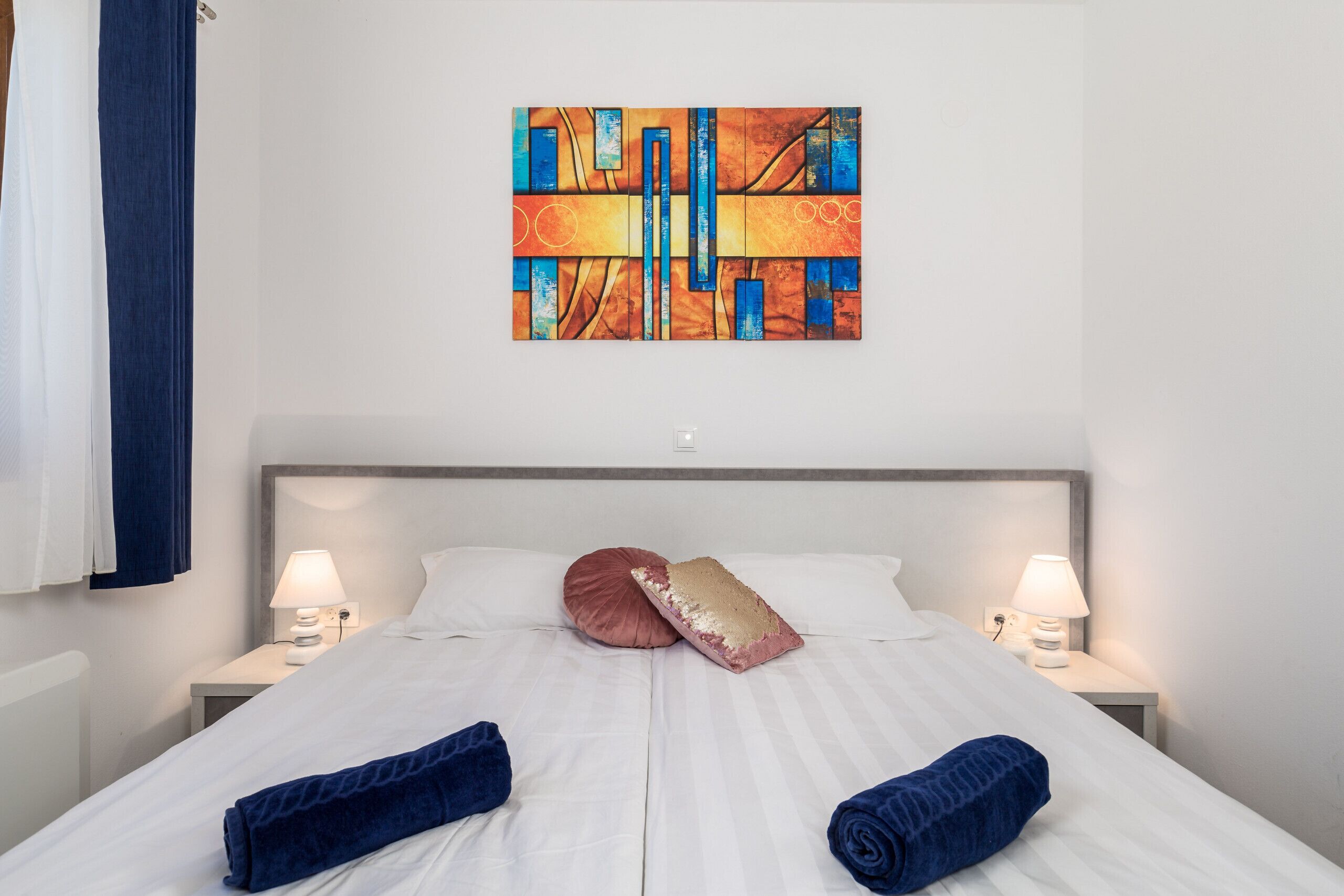 Una habitació d'hotel amb un llit gran, dues làmpades de nit, un capçal de llit, una tovallola blava, un coixí rosa i un quadre abstracte i colorit a la paret.