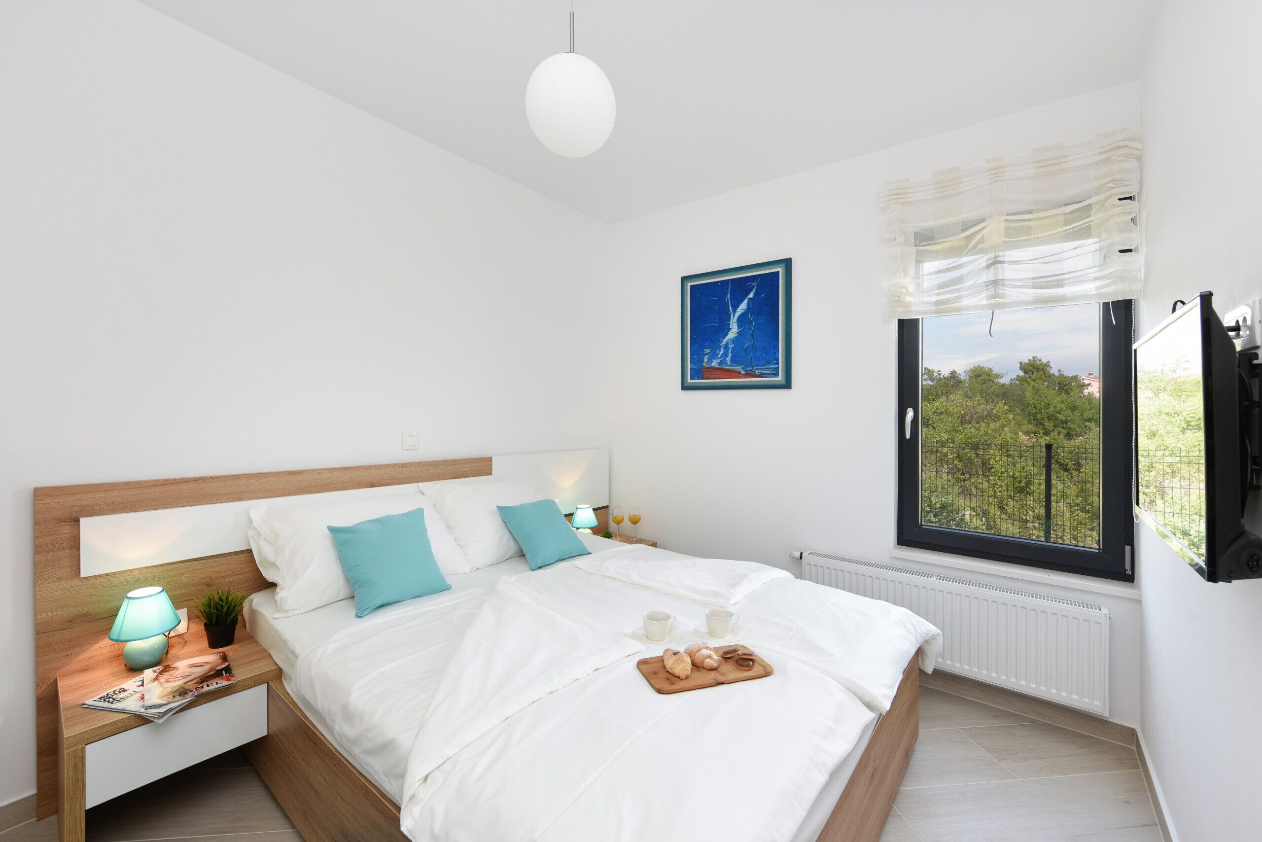 A modern bedroom with a large bed, a bedside table with a lamp, a window with a view, and a framed picture on the wall.