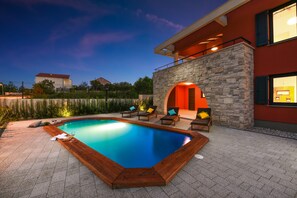 Una piscina con muelle de madera y sillones de descanso, rodeada por un muro de piedra y un edificio rojo con balcón.