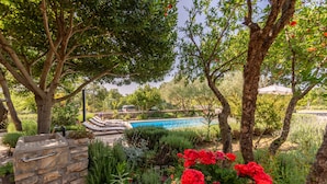 A pool area with stone walls and a stone grill, surrounded by lush greenery and flowering plants. - Villa Krasa (Promina)