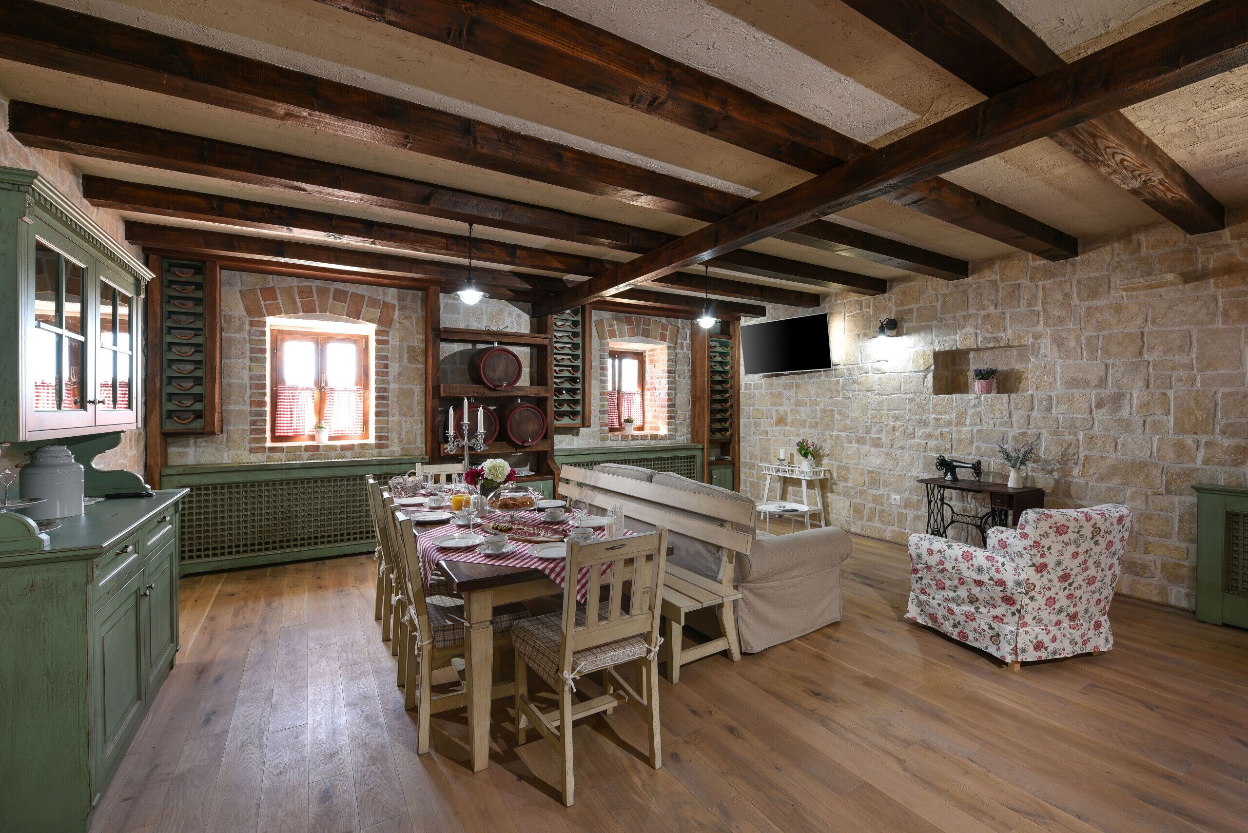 Un comedor rústico con una mesa de madera preparada para cuatro comensales, rodeada de sillas. Se observa una pared de piedra y un televisor montado en ella al fondo.