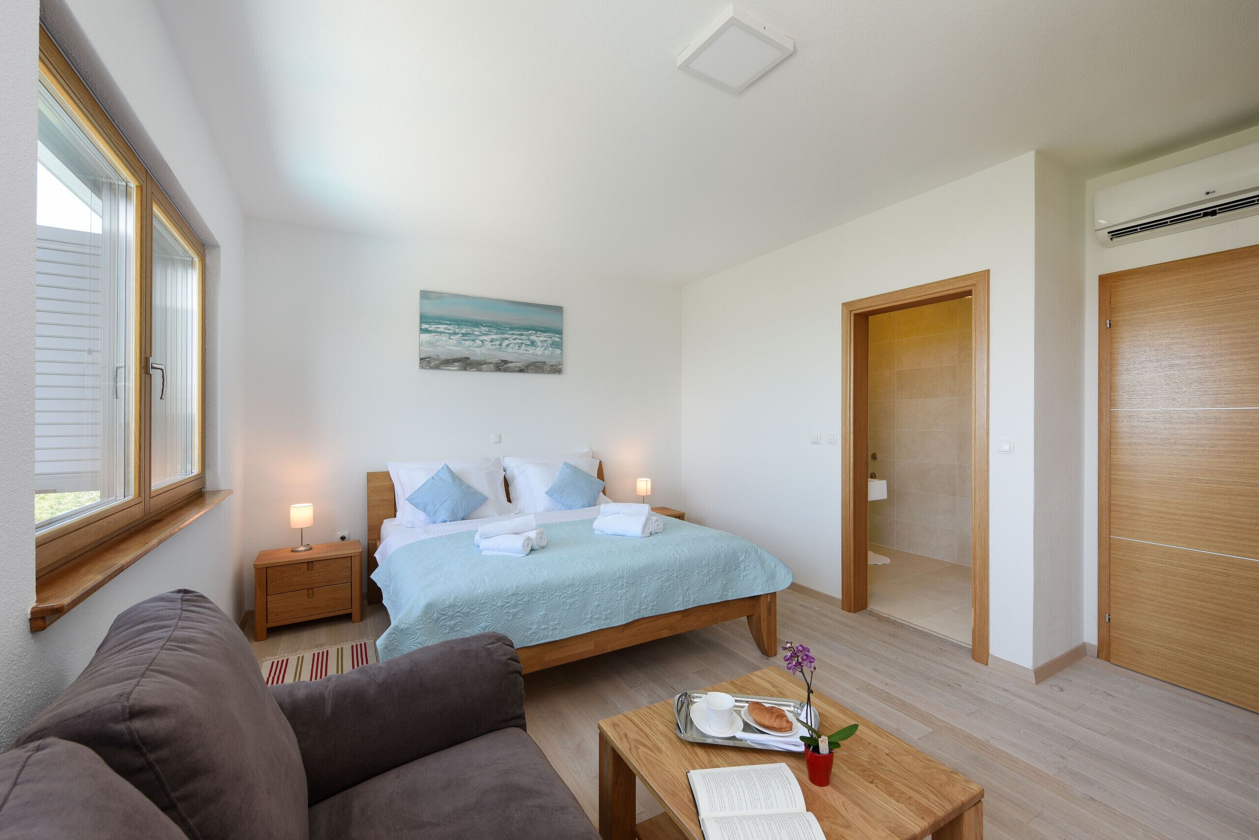 A bedroom with a bed, bedside tables, a nightstand, a window, a painting, a wooden door, and a wooden floor.