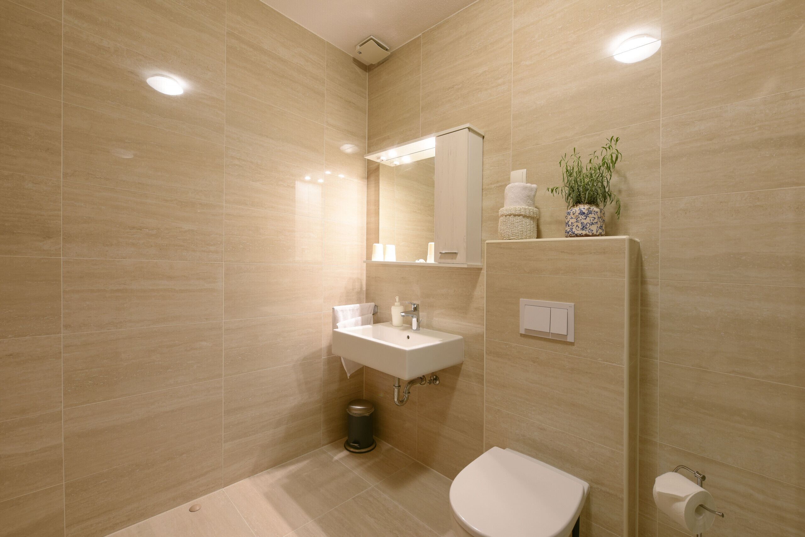 A modern bathroom with a beige tiled wall, a white sink, a mirror, a toilet, and a trash bin.