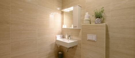 A modern bathroom with a beige tiled wall, a white sink, a mirror, a toilet, and a trash bin.