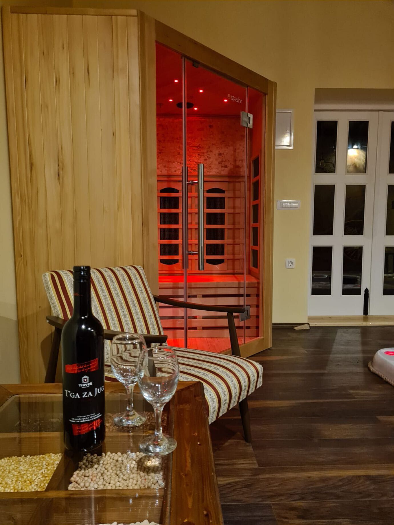 Quarto com estrutura de madeira, porta deslizante vermelha, poltrona estampada e mesa com garrafa de vinho e copos.