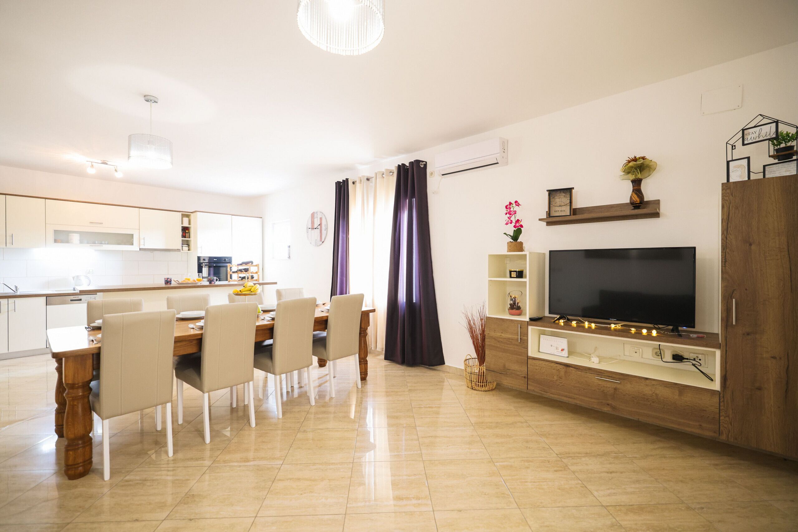 A modern kitchen with white cabinets, a dining area with a wooden table and chairs, and a flat-screen TV on a wooden stand.