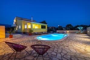A stone-paved outdoor area with a swimming pool, lounge chairs, and a house with illuminated windows. - Villa Didov Dvor (Posedarje)