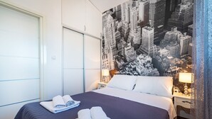 A hotel room with a large bed, two rolled towels, a cityscape mural on the wall, a white wardrobe, and a nightstand with a lamp. - Villa Didov Dvor (Posedarje)