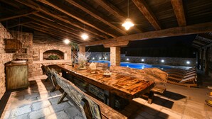 An outdoor dining area with a rustic wooden table and benches, stone walls, and a pool in the background. - Villa Quince (Policnik)
