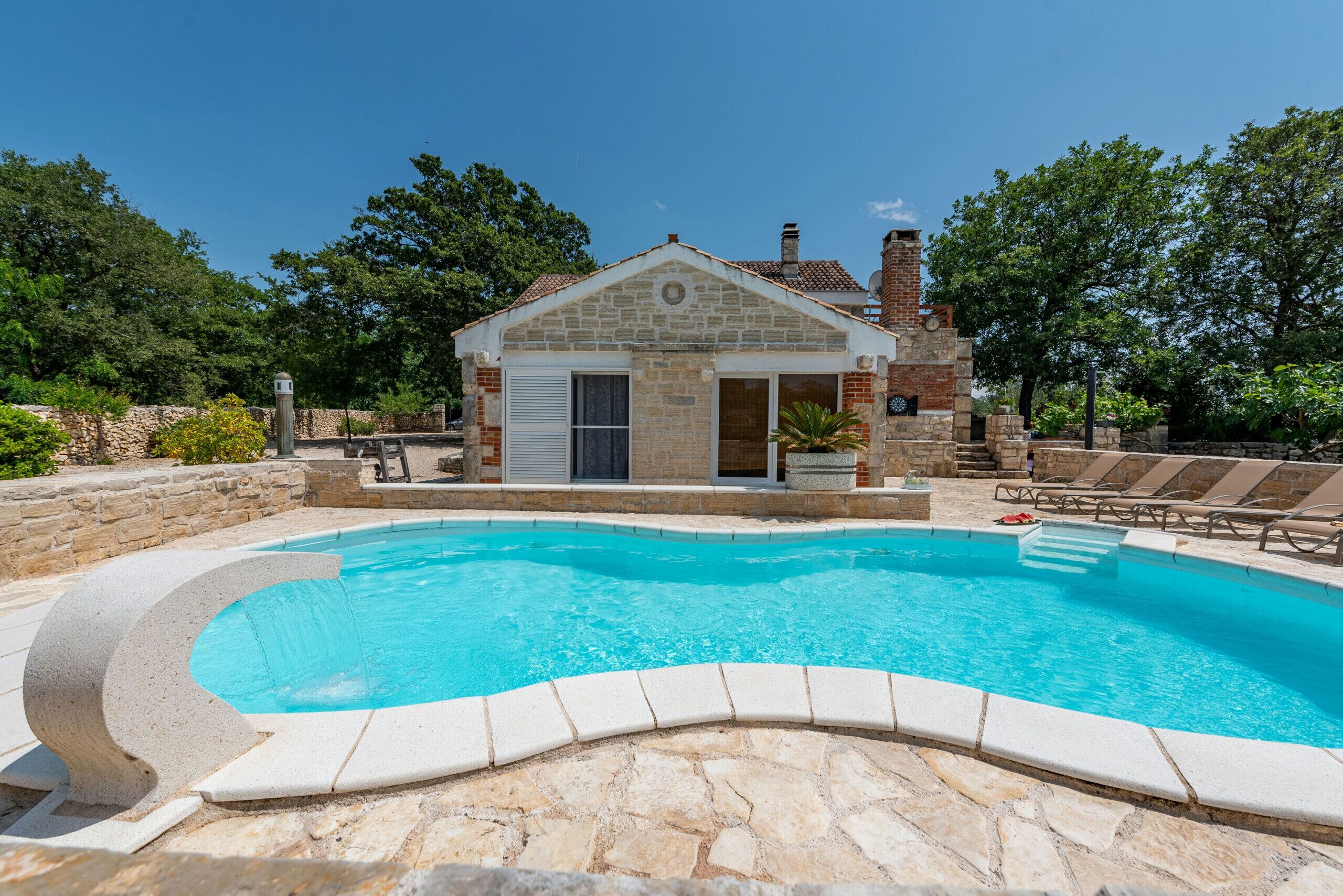 Uma piscina com borda de pedra, um pequeno edifício de pedra com uma porta e uma chaminé de tijolos.