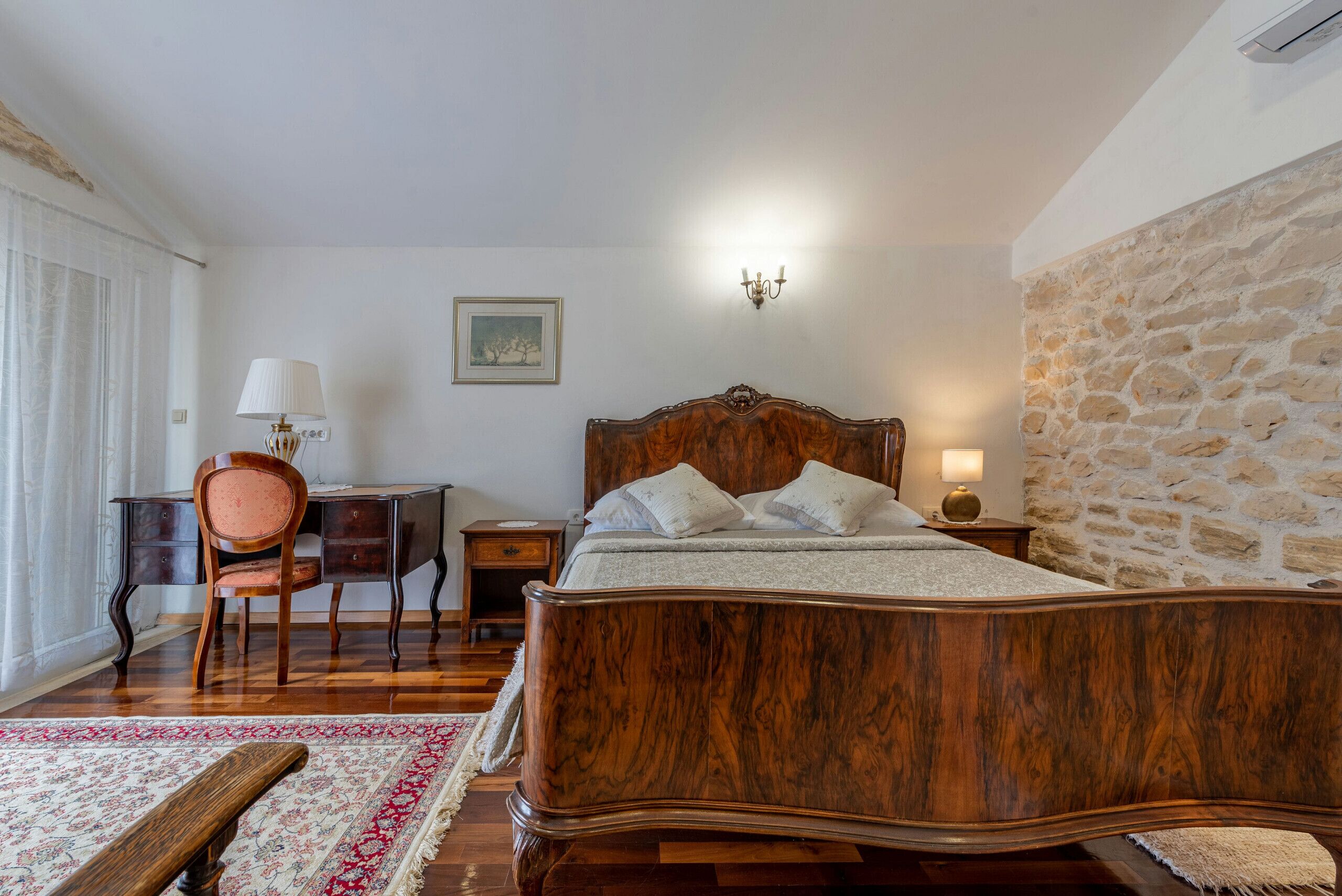 A bedroom with a wooden bed, a desk, a chair, and a stone wall.
