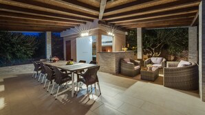 Un área de comedor al aire libre con una mesa larga y sillas, rodeada de mobiliario de mimbre y un muro de piedra.