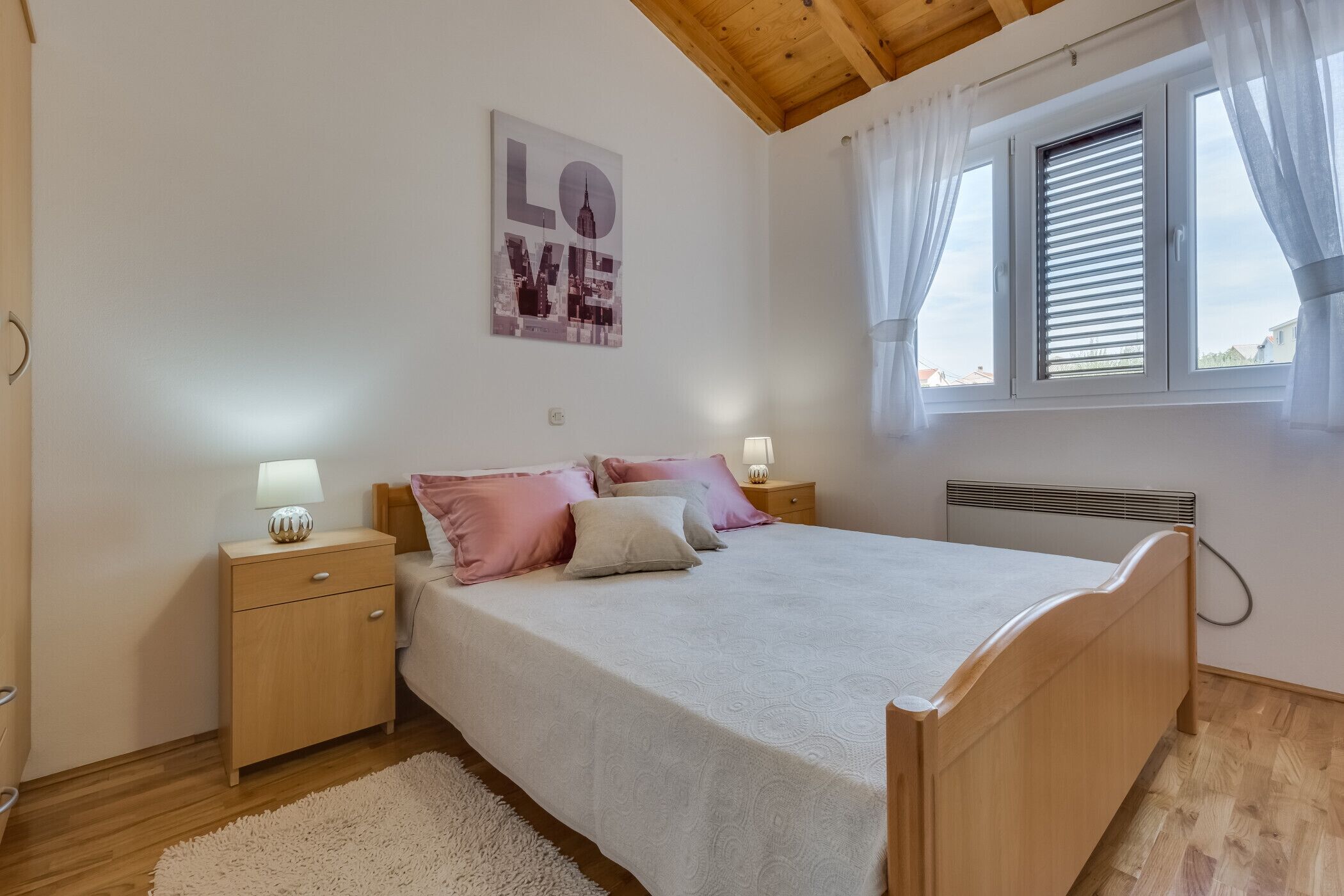 Een slaapkamer met een houten bed, een nachtkastje met een lamp, een raam met zonwering en een poster van een stadsgezicht aan de muur.