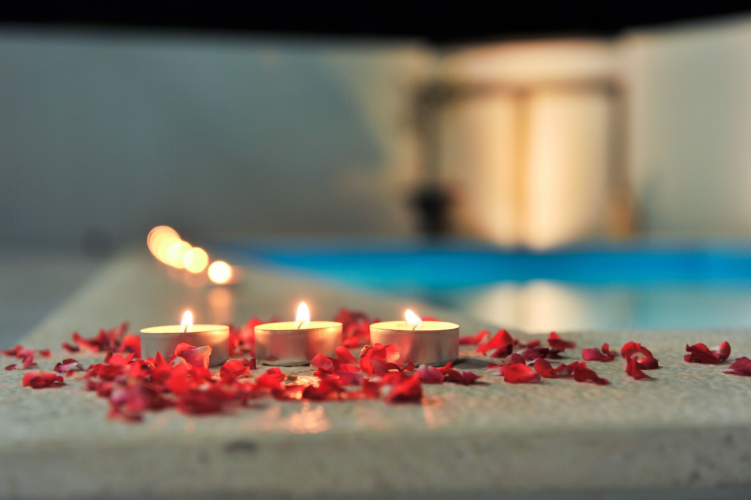 Three lit candles on a stone surface with red petals scattered around.