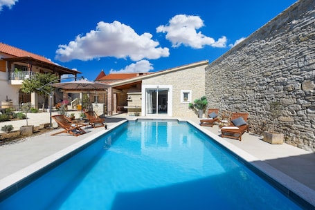 A swimming pool with lounge chairs, a stone wall, and a building with a tiled roof.