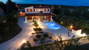 Una casa de dos pisos con piscina, mobiliario exterior y un patio de piedra.
