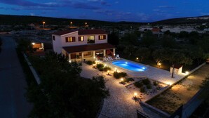 Una casa de dos pisos con piscina, rodeada por un sendero de piedra y mobiliario de exterior.