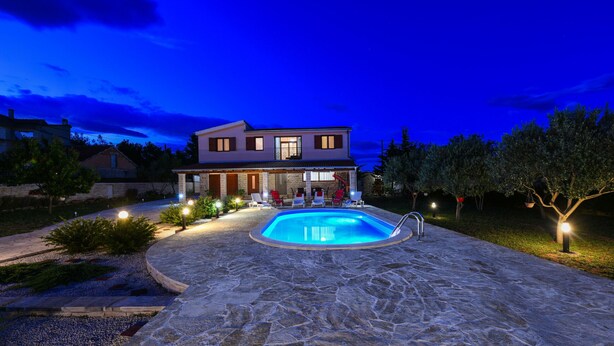 Una casa de dos pisos con piscina, patio de piedra y zona de estar exterior.