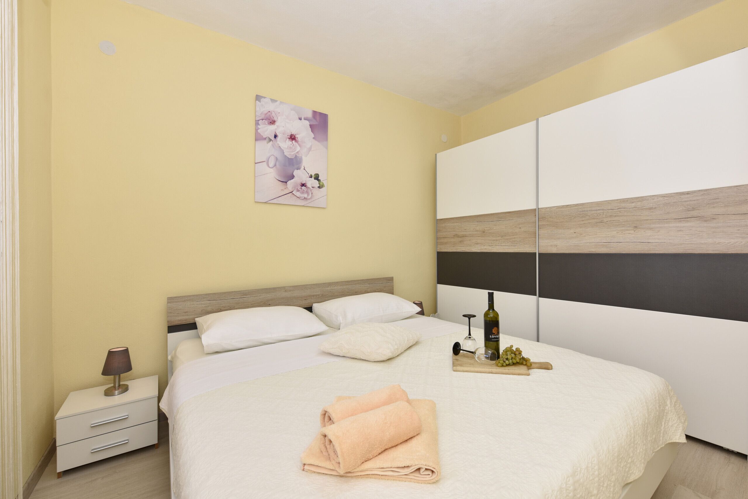A neatly made bed with a white comforter and a folded towel, a bedside table with a lamp, a framed picture on the wall, and a wooden headboard.