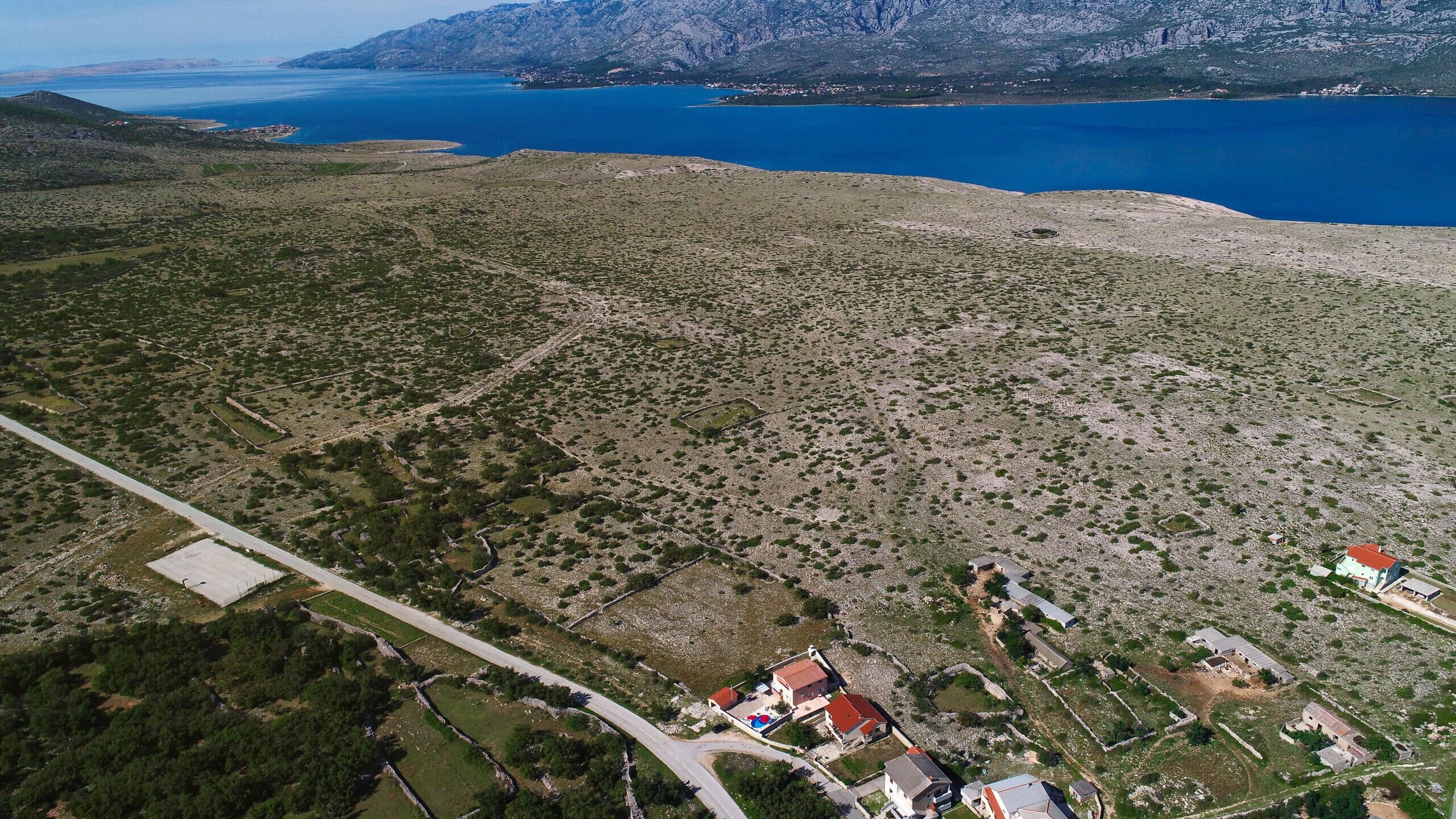 An aerial view of a rural area with scattered buildings, a road, and a large body of water in the background.