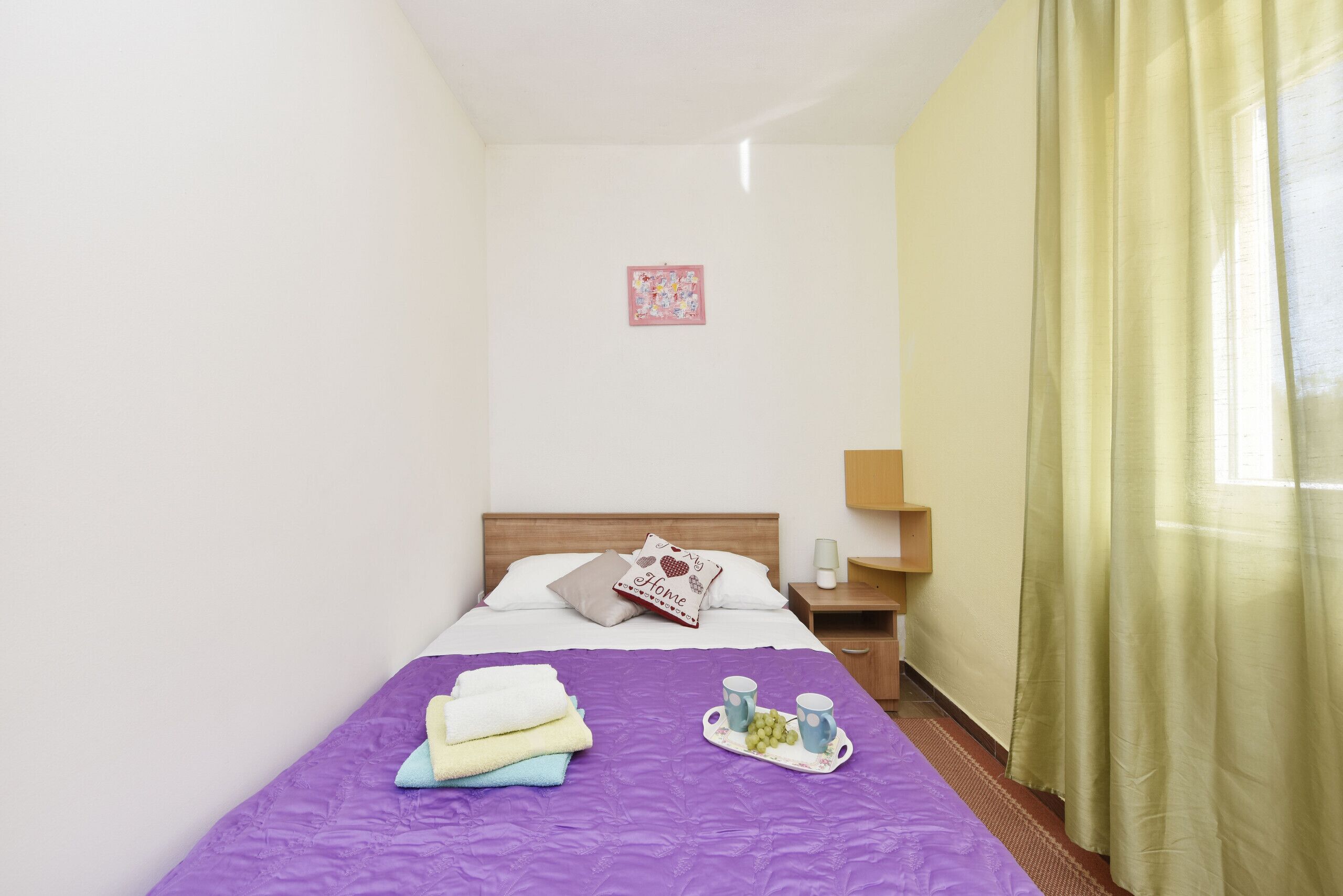 A neatly made bed with a purple bedspread, a tray with cups and grapes, and a pillow with a heart design.