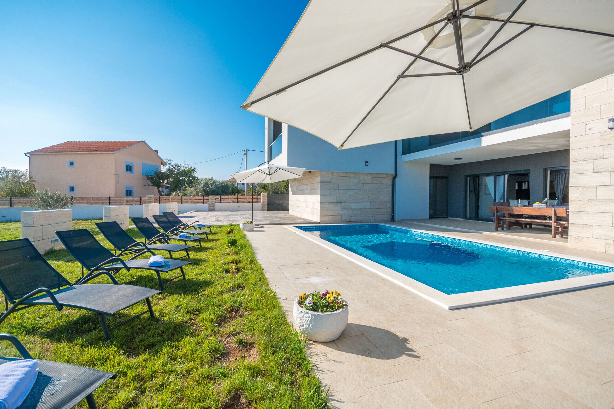 A modern outdoor pool area with lounge chairs and umbrellas, a clear blue sky, and a building in the background.