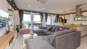A modern living room with a grey sofa, a glass coffee table, and a wooden side table. There is a kitchen area with a stove and a dining table. - Villa F&A (Sveti Filip i Jakov)
