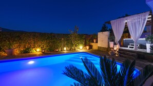 Una piscina iluminada con luz azul durante la noche, rodeada de plantas en macetas y un pérgola con cortinas blancas.