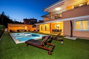 Una casa de dos pisos con piscina, mobiliario exterior y un patio bien iluminado.