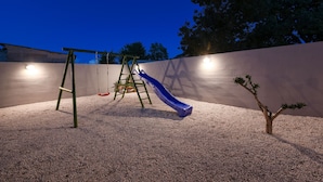 Ein Spielplatz mit Rutsche und Schaukeln, umgeben von einer Mauer und einem kleinen Baum.
