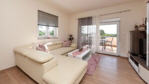 Ein Wohnzimmer mit einer Couch, einem Fernseher, einem Balkon mit Tisch und Stühlen sowie einer Schiebeglastür.