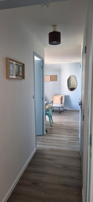 A hallway with a wooden floor, a door, a chair, a table, and a mirror on the wall.