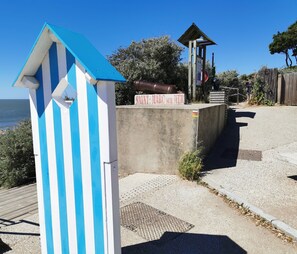 Una estructura frente a la playa con techo azul y blanco a rayas, pared de hormigón y un pequeño edificio de madera con un letrero.