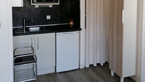 Una cocina compacta con armarios blancos, encimera negra, microondas, pileta y un refrigerador pequeño, además de un carrito metálico.