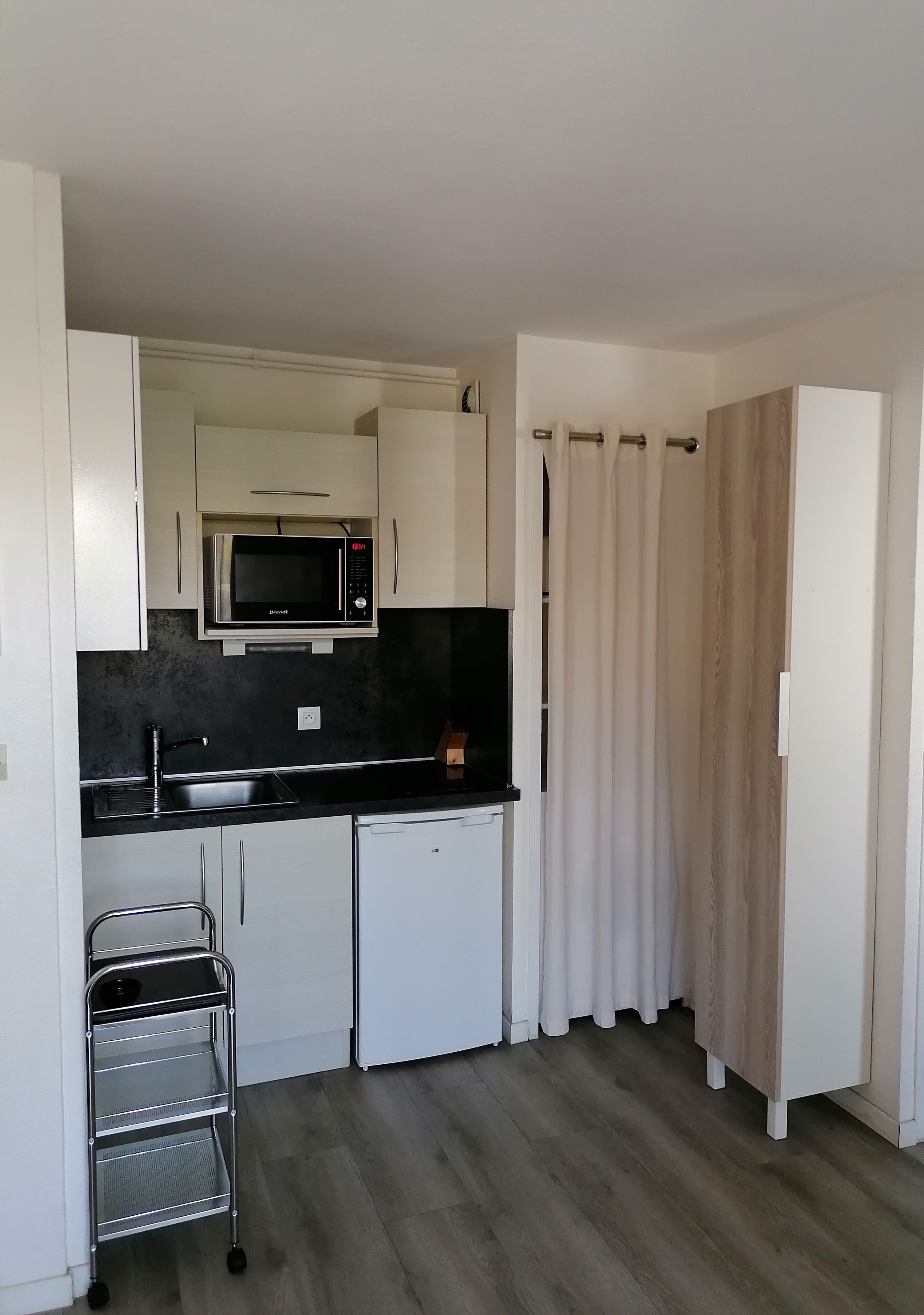 A compact kitchen with white cabinets, a black countertop, a microwave, a sink, a small refrigerator, and a metal cart.