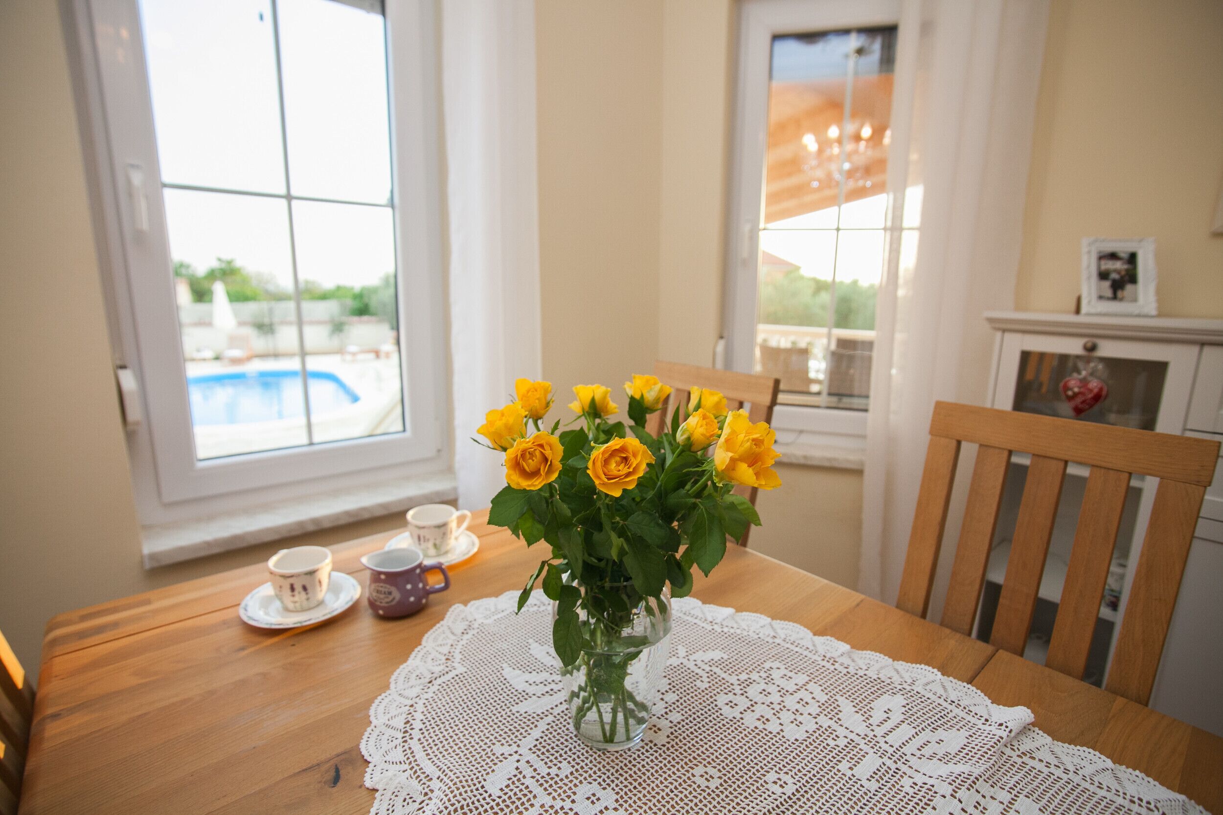 A dining table with a clear view of a swimming pool through a window, a vase with yellow flowers, and a wooden chair.