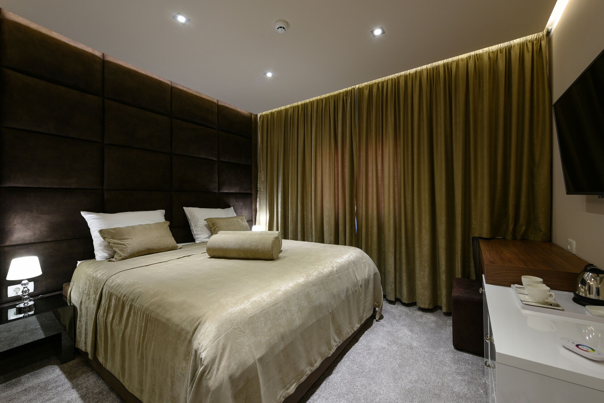 A modern hotel room with a large bed, a bedside table with a lamp, a desk with a chair, and a large window with curtains.