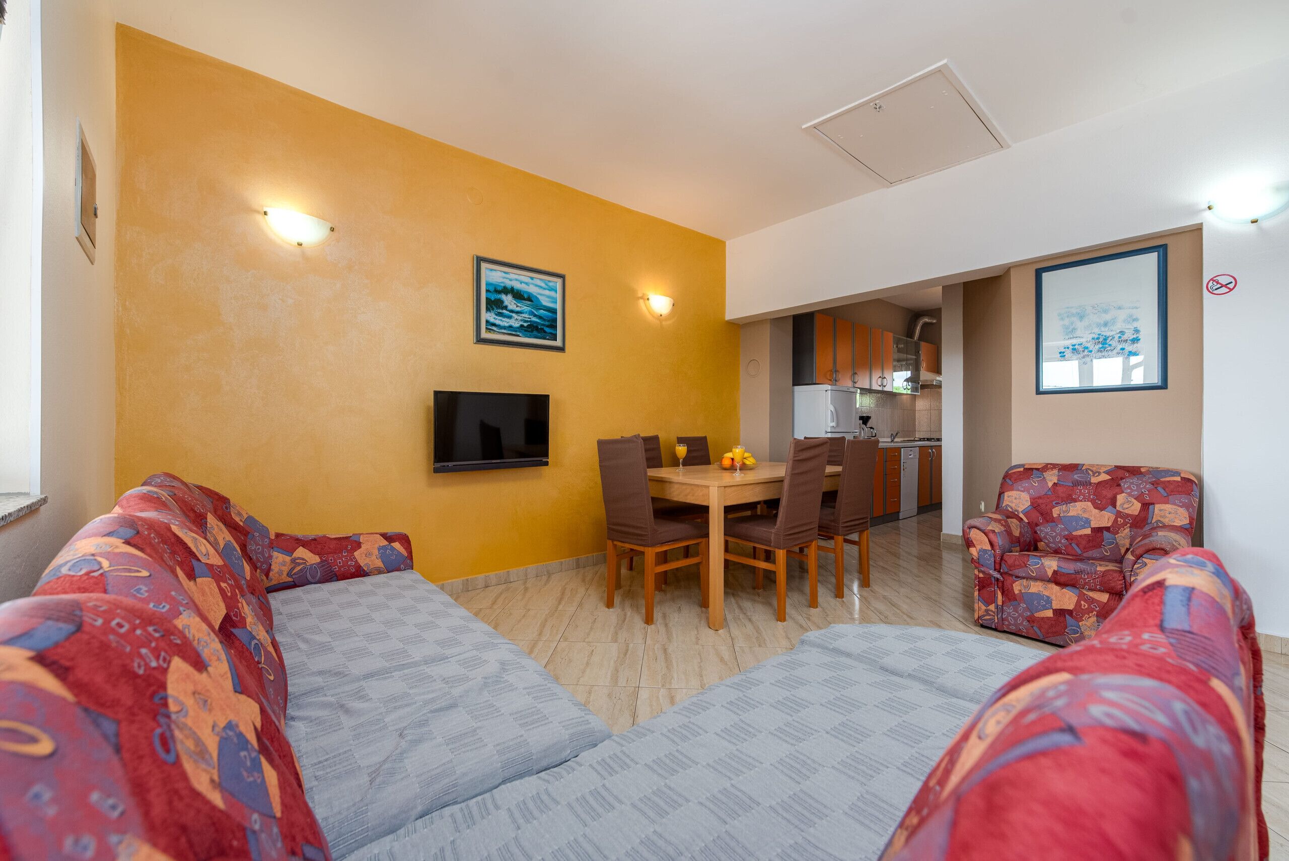A living room with a sofa, a dining table with chairs, a TV mounted on the wall, and a kitchen in the background.