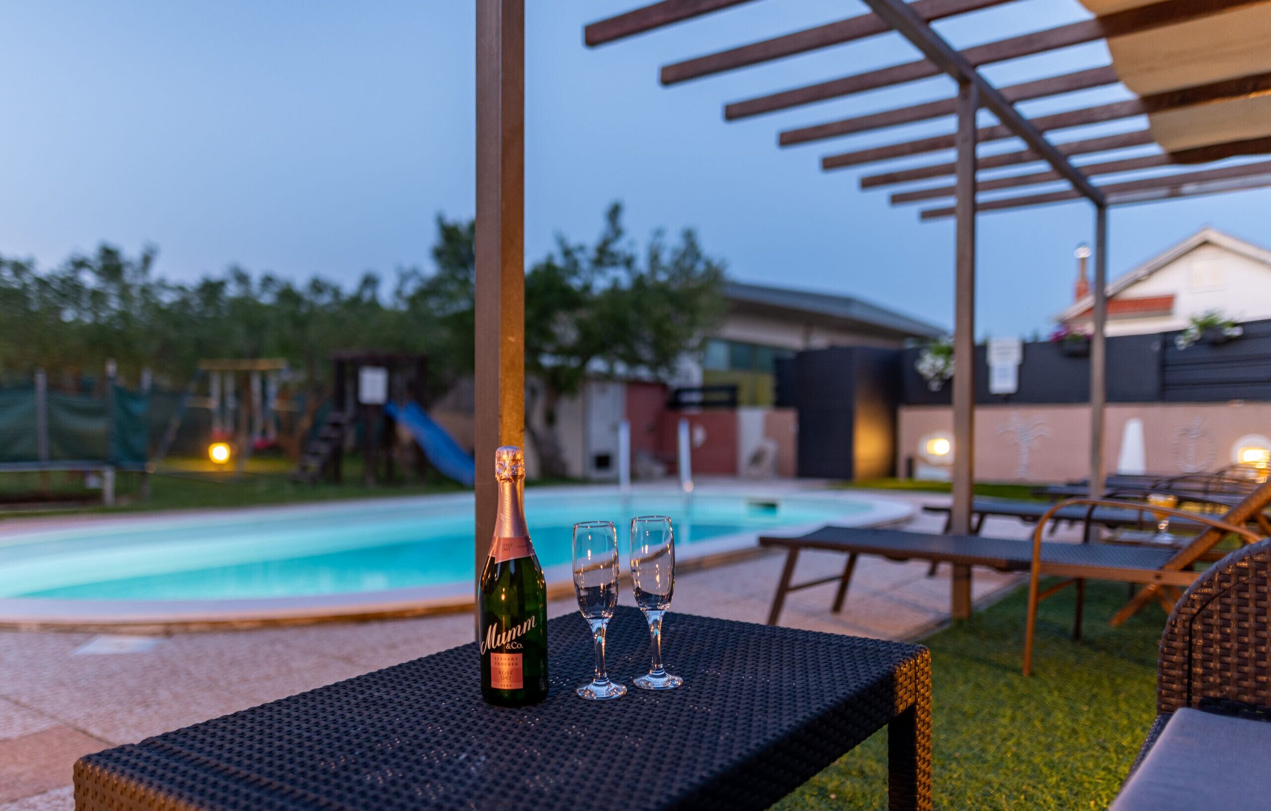A poolside setting with a bottle of champagne and two glasses on a table, surrounded by lounge chairs and a pergola.