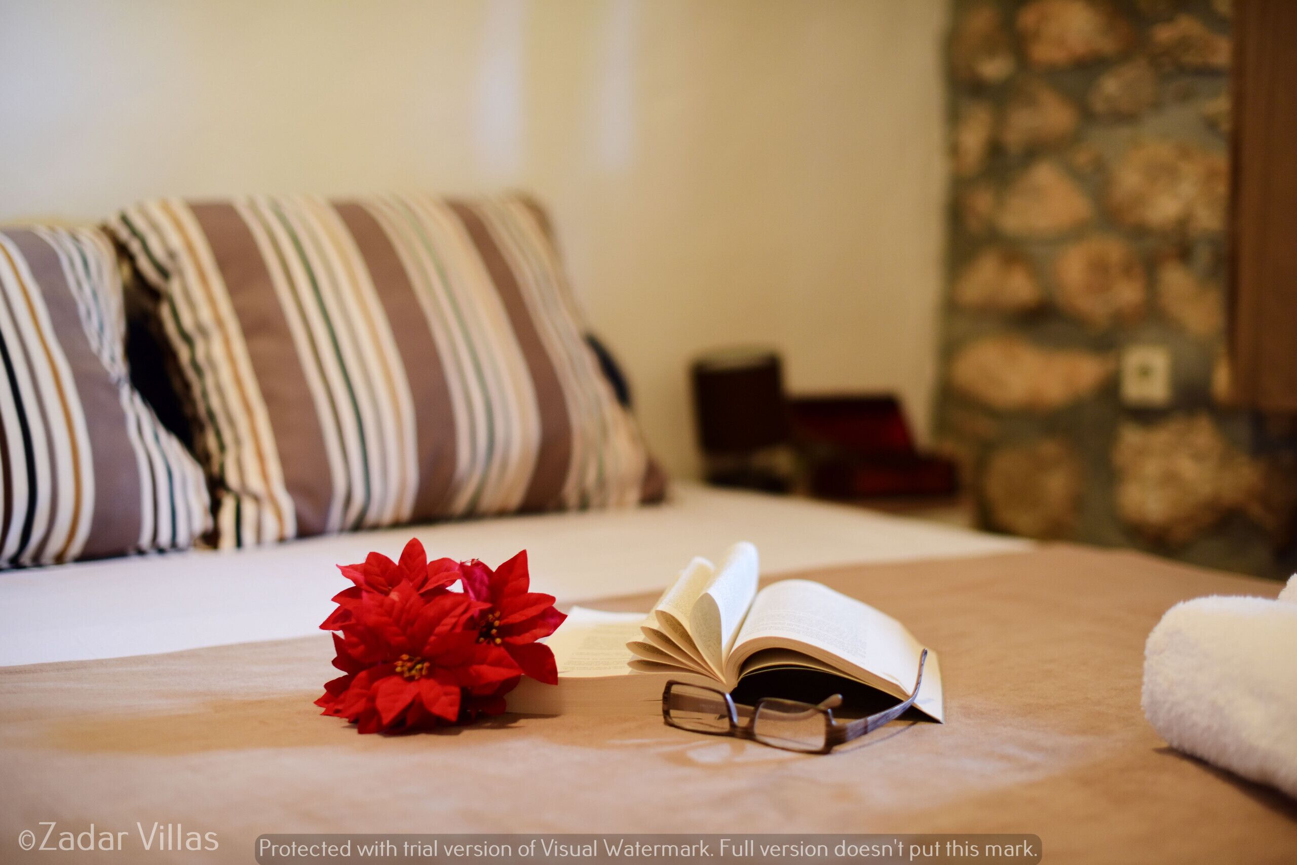 Un lit bien fait, orné d’oreillers à rayures, une fleur rouge et un livre ouvert, avec des lunettes posées à côté.