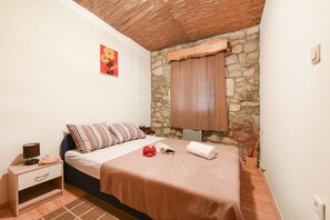 Een slaapkamer met een bed, een nachtkastje, een lamp, een raam met gordijn en een stenen muur.