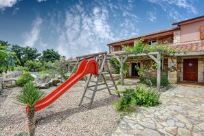 Een wooneiland met een stenen terras, een rode glijbaan en een houten pergola met klimplanten.
