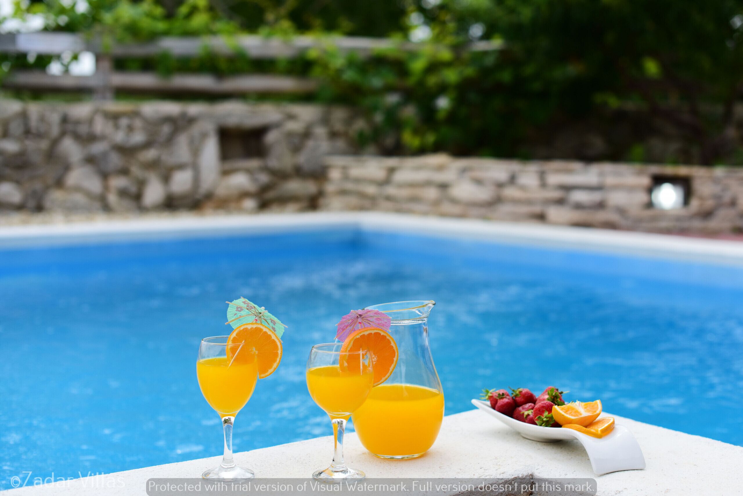 A poolside setting with a table holding two glasses of orange juice and a pitcher, accompanied by a plate of strawberries and oranges.