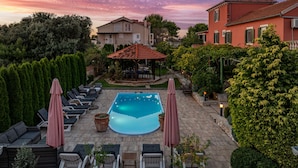 A swimming pool with lounge chairs and umbrellas, surrounded by greenery and a building with a red roof. - Villa Stocco (Preko)